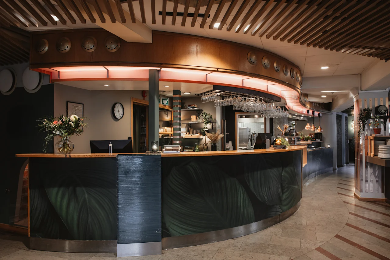A stylish and welcoming reception or bar area at Home Hotel Uppsala, featuring a curved counter with a leafy design and hanging glasses.