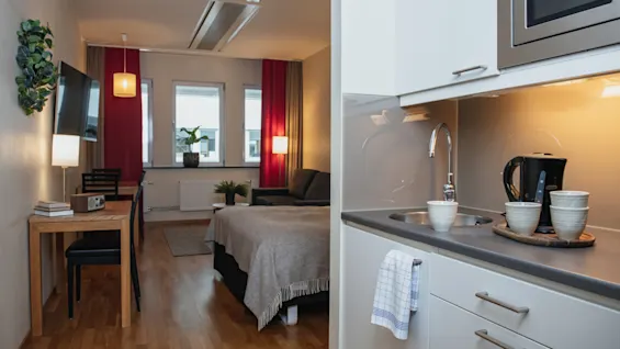 A small studio apartment with a kitchenette, featuring a sink and cups on a tray. A bed and dining table with two chairs are in the well-lit living area.