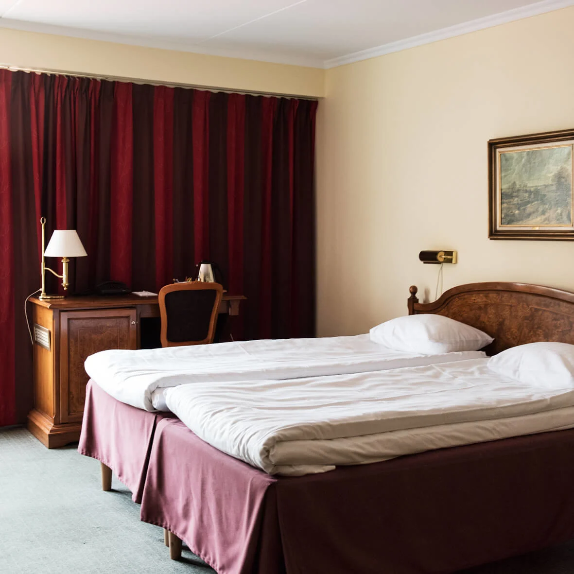 En dobbeltseng med hvitt sengetøy og en trehodegavl står på et hotellrom. Mørkerøde gardiner dekker vinduet, og et skrivebord med en lampe er i nærheten.