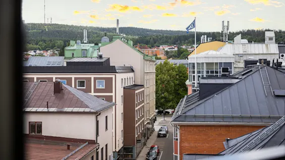 Stadsbild med flera hustak, en flagga som vajar, mot en delvis molnig himmel med träd och kullar i bakgrunden.