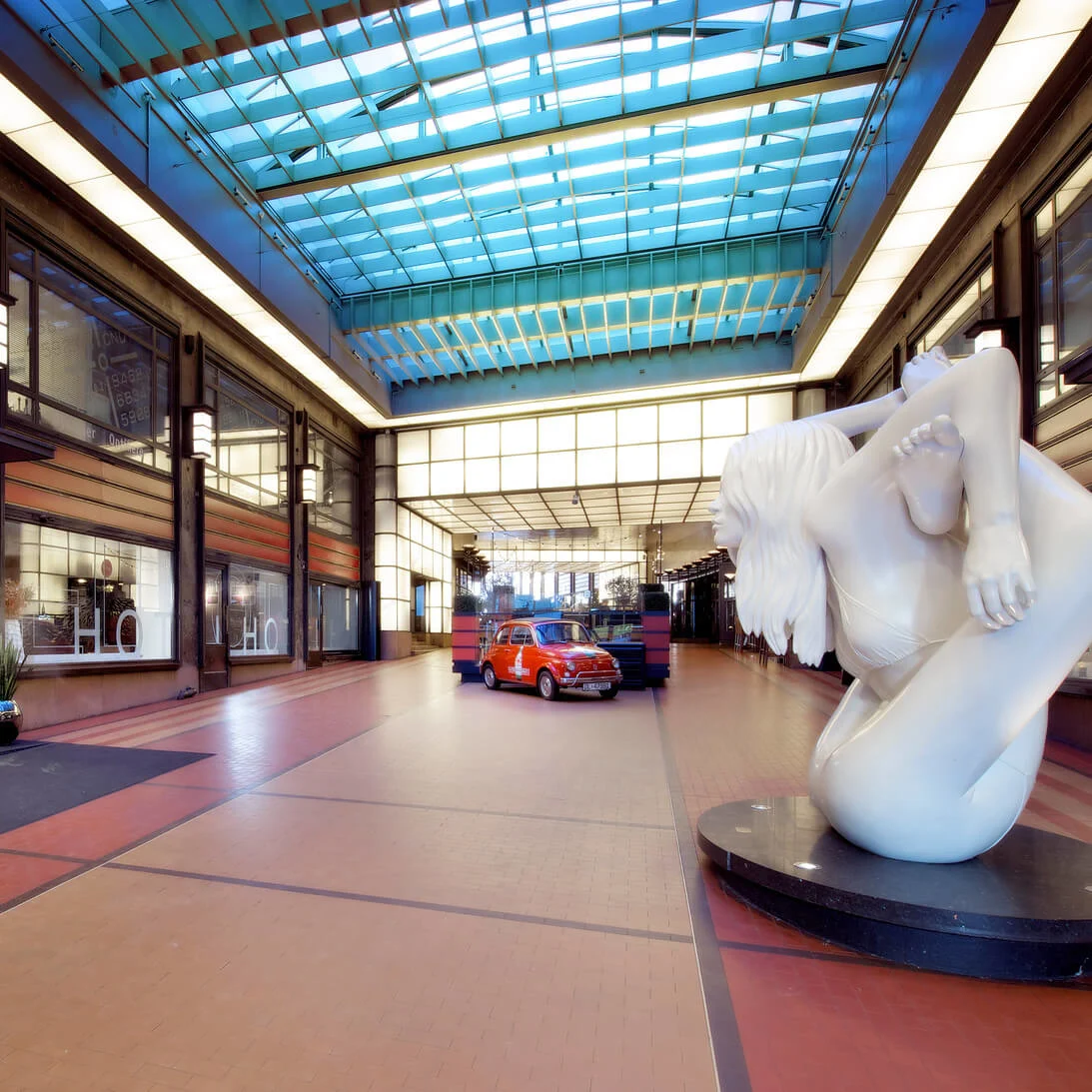 En skulpturert figur sitter ved siden av parkerte biler under et blåaktig overlys i et industrielt innendørsrom.
