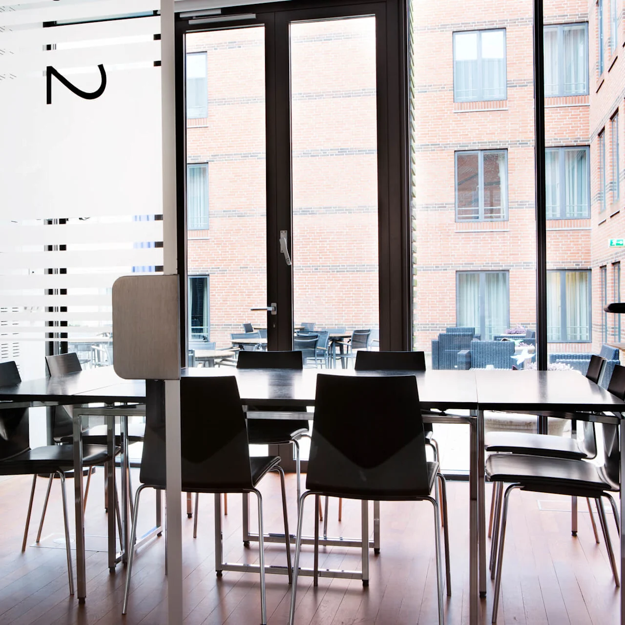  A bright meeting room with glass walls and a view of a courtyard. Minimalist-style tables and chairs are placed on a wooden floor, with the number "2" marked on the glass to identify the room.