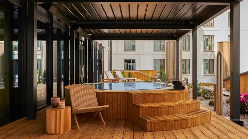 A wooden hot tub sits on a deck under a modern pergola. Nearby, chairs and tables create a relaxing patio setting, surrounded by contemporary apartment buildings with large windows.