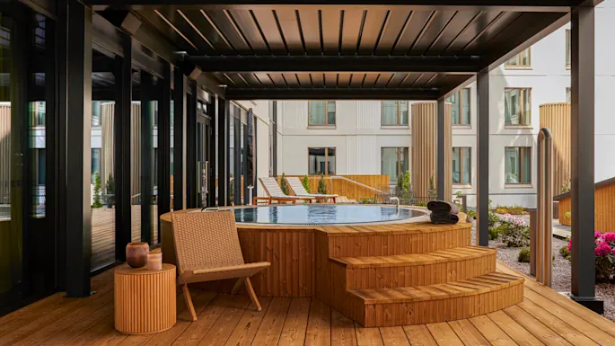 Et boblebad i tre står på en terrasse under en moderne pergola. I nærheten skaper stoler og bord en avslappende utemiljø, omgitt av moderne leilighetsbygg med store vinduer.