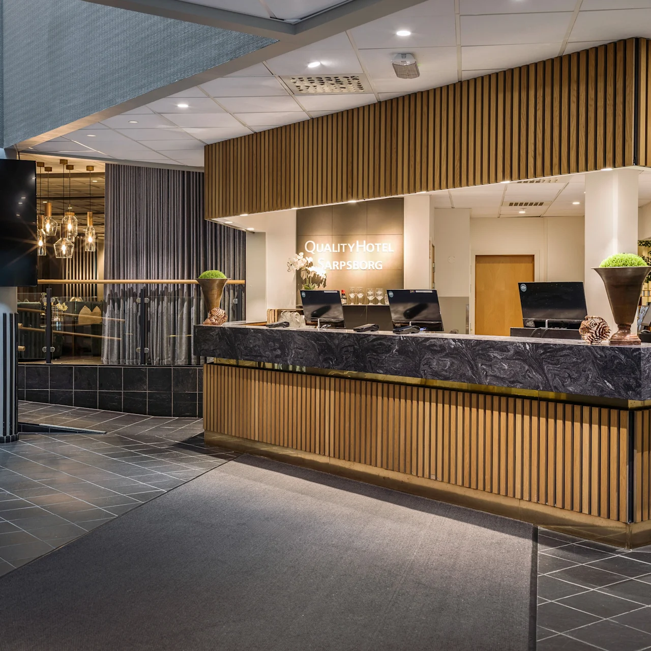 Reception desk with computers and decorative items sits in a modern hotel lobby. Behind the desk, a sign reads "QUALITY HOTEL SARPSBORG" with stylish lighting and tiled flooring around.