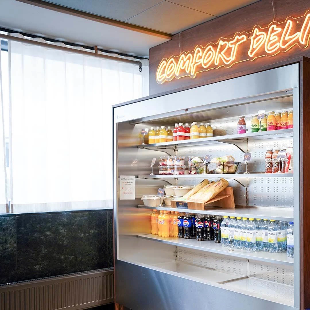 A refrigerator with beverages and snacks in a brightly lit deli; neon sign "COMFORT DELI" overhead.