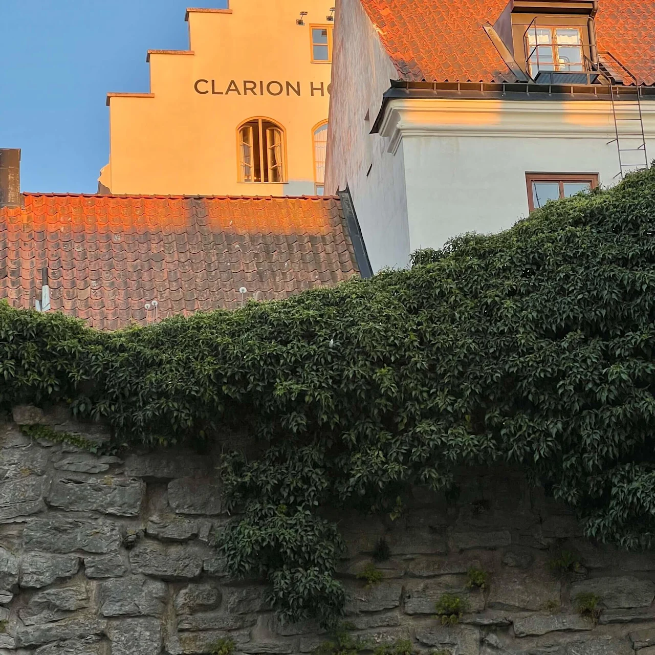 En bygning med rødt tegltag og trappegavl er oplyst af solnedgang, set over en stenmuren dækket af frodigt grønt. Tekst: "CLARION HO".