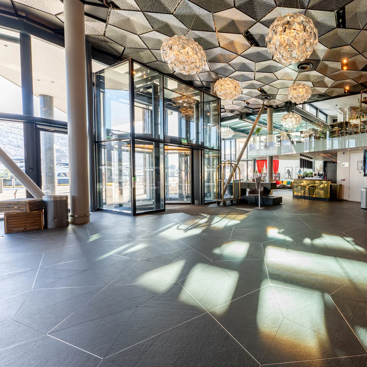 Stor glaselevator reflekterer sollys i en moderne hotellobby med dekorativ belysning og receptionsskranker. Tekst inkluderer "SOCIAL," "Check today's weather," og "CLARION HOTEL" ved indgangen.