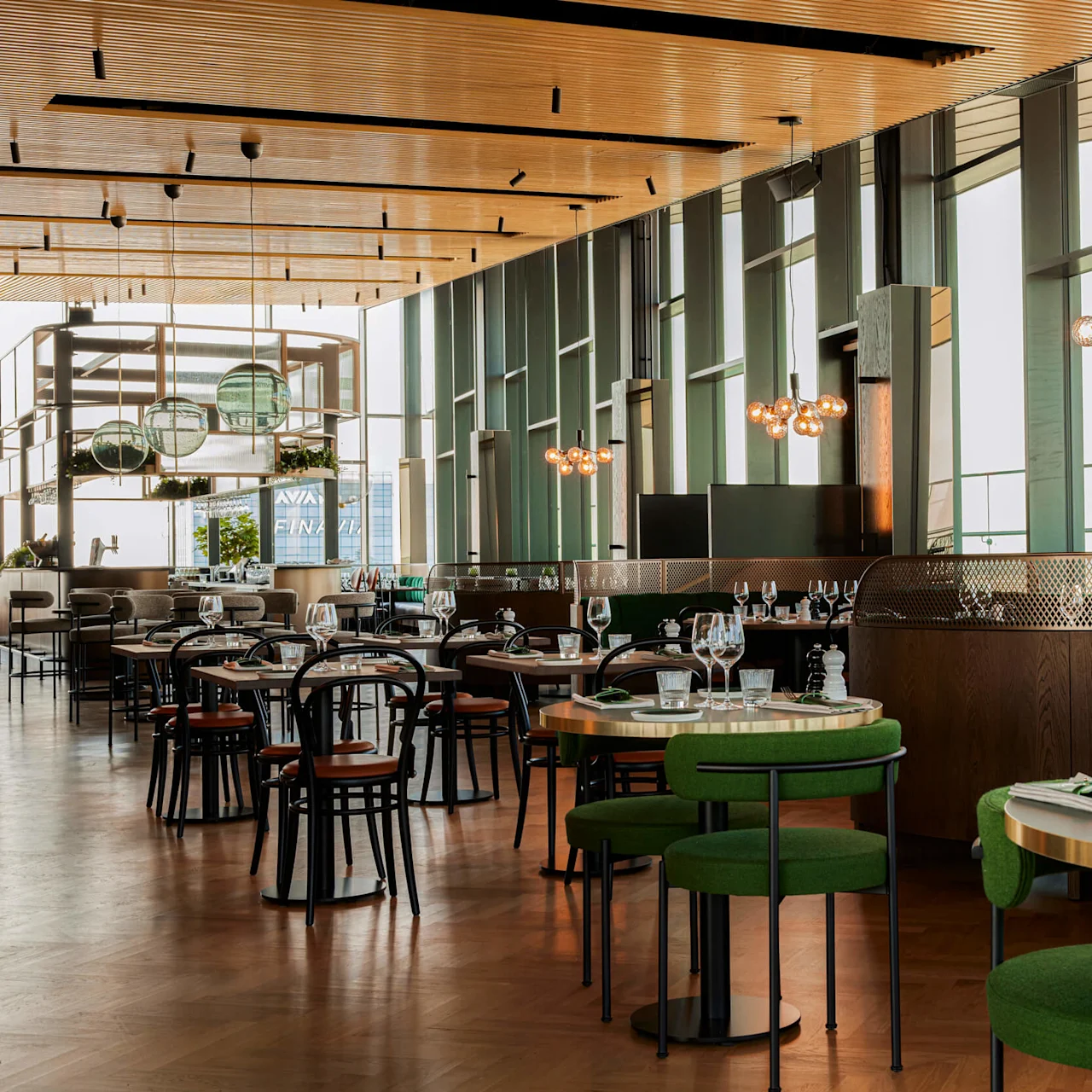 Modern restaurant setting; tables with green chairs and neatly set with glasses and cutlery. Large windows let in natural light, with pendant lights hanging from the ceiling.