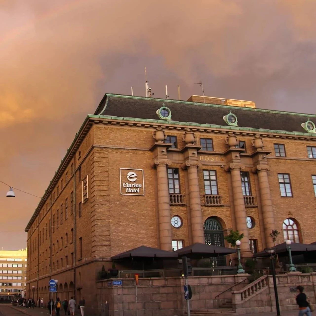 Suuri tiilirakennus, jossa on kyltti "Clarion Hotel", seisoo sateenkaaren alla auringonlaskun taivalla. Ympärillä on katulamppuja ja ihmisiä kävelemässä alapuolella.