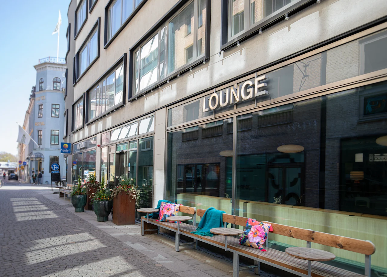 Et indbydende udendørs loungeområde med bænke og planter foran en bygning med store vinduer. Skiltet siger LOUNGE.