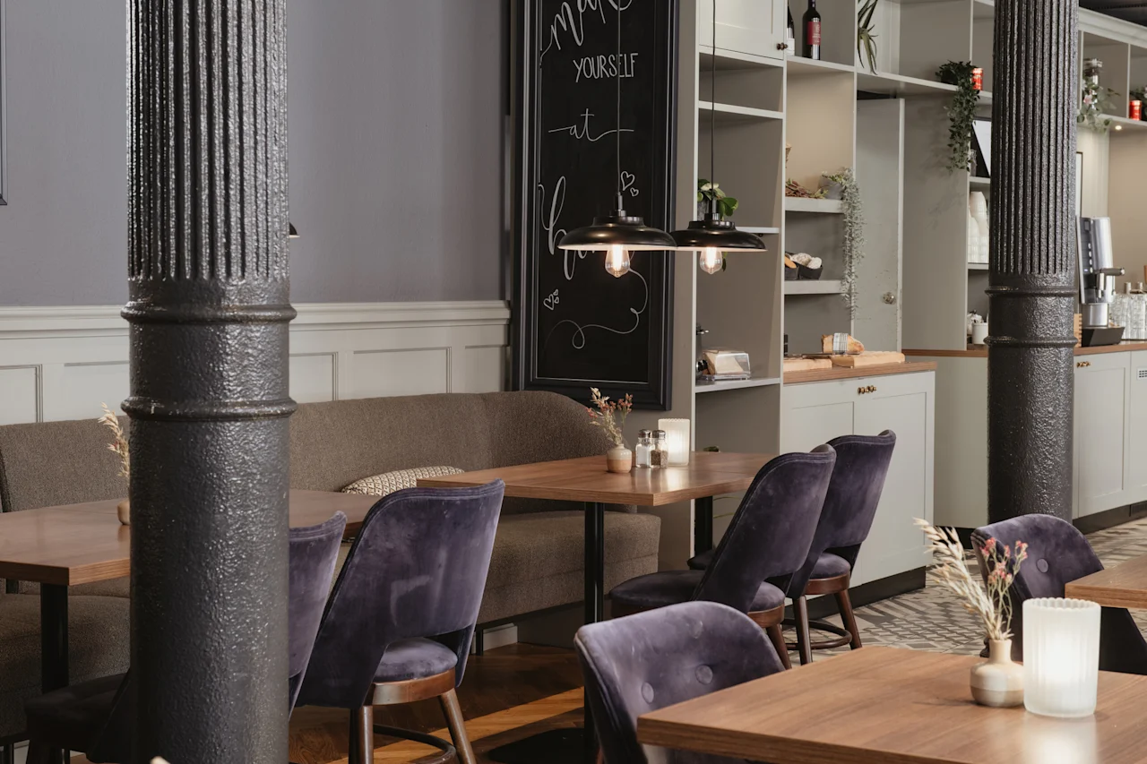 Et koselig kaféinteriør med trebord, lilla fløyelsstoler og et krittskilt som sier 'make yourself at home'.