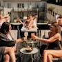Folk kopplar av på solstolar och njuter av drinkar på en solig takterrass omgiven av byggnader. Två kvinnor i förgrunden skålar med färgglada cocktails, omgivna av små bord och växter.