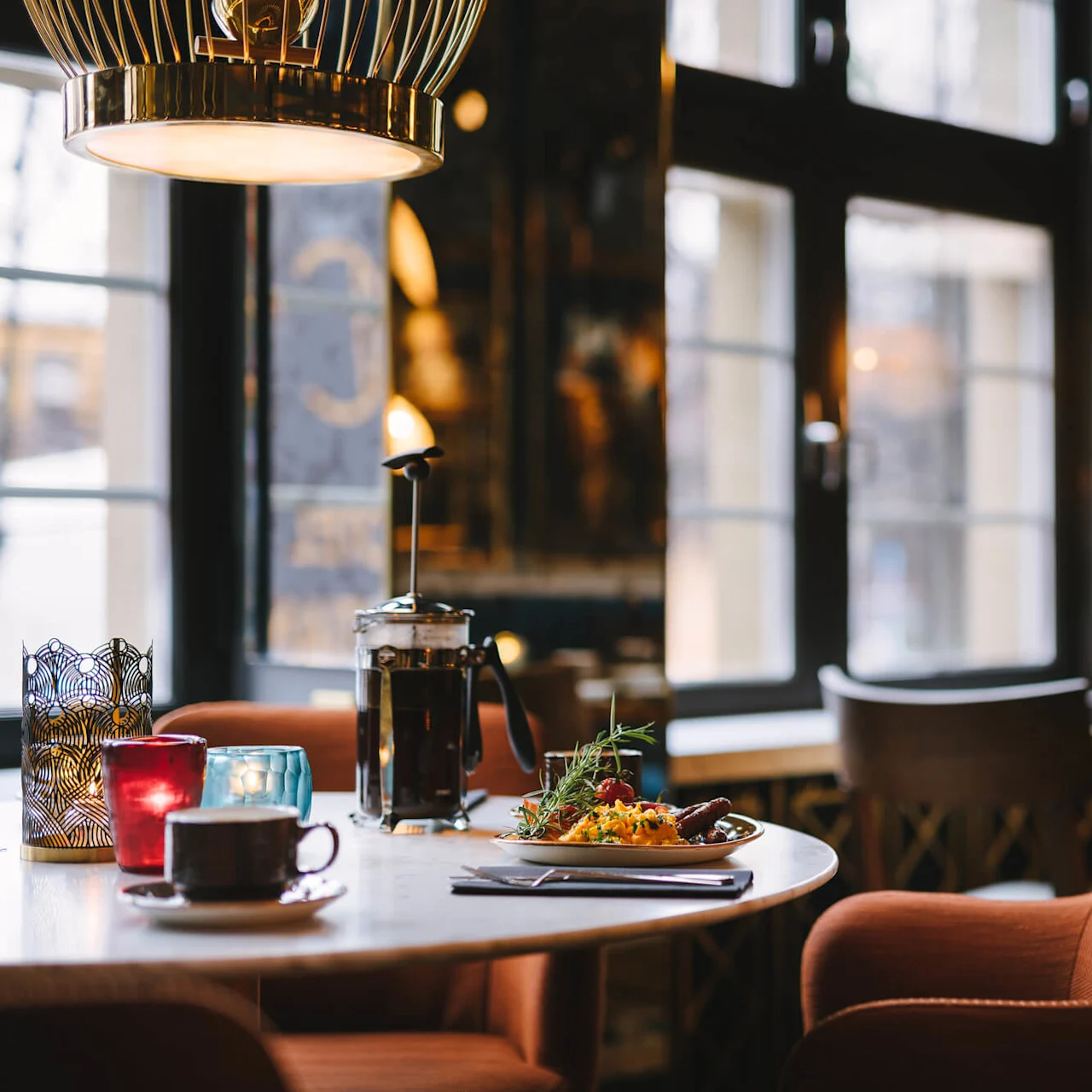 A table in a cozy café holds a French press, a filled plate, and a coffee cup. Warm lighting filters through large windows, with orange chairs surrounding the scene.