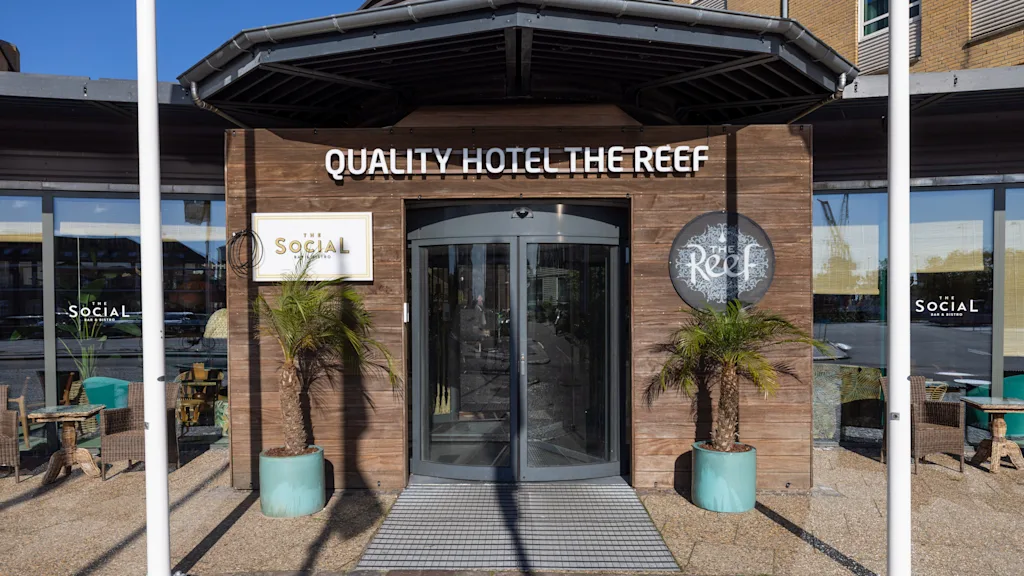 En ingång av glas märkt "QUALITY HOTEL THE REEF" med sido-skyltar "SociAL" och "Reef"; omgiven av växter i krukor på en skuggig uteplats med sittplatser utomhus.