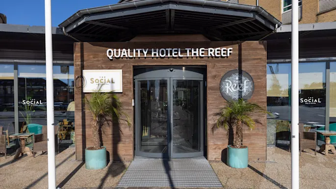 En glassdør merket "QUALITY HOTEL THE REEF" med skilt på sidene "SOciAL" og "Reef"; omgitt av planter i potter på en skyggefull uteplass med uteservering.