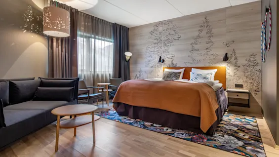 A bed with orange bedding is placed against a wall featuring tree illustrations. Nearby, a gray sofa and round table enhance the cozy hotel room atmosphere.