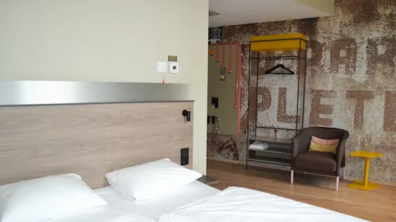 A neatly made bed with white linens is in the foreground. A chair, yellow table, and hanger are in the background. The wall has the partial text "PLET" printed on it. 
