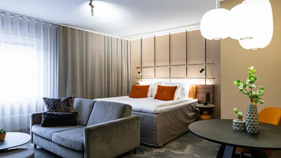 Et stilfuldt hotelværelse har en pænt redt dobbeltseng med orange puder, placeret ved siden af en grå sofa. Et moderne rundt bord med vaser er i nærheden under blød belysning.