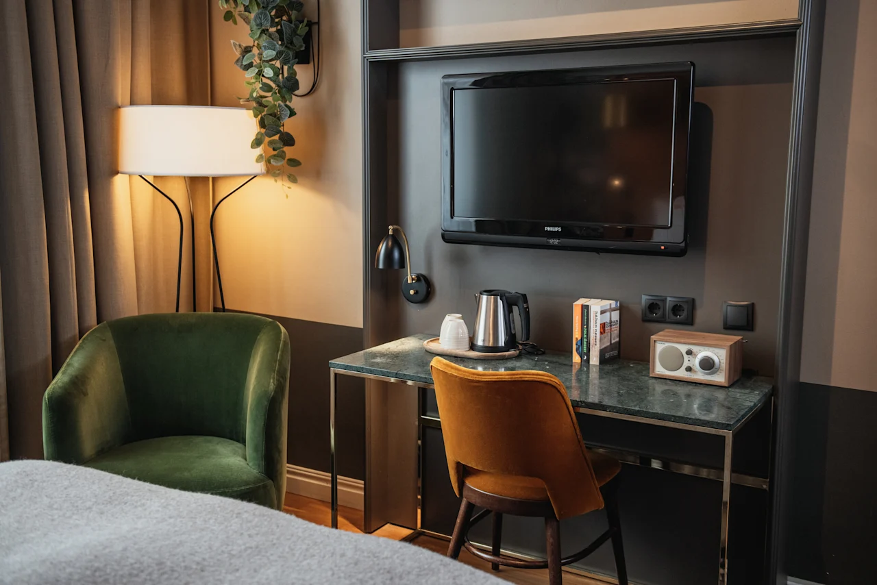 Et velutstyrt hotellrom med et funksjonelt skrivebordsområde med TV, vannkoker og radio, supplert med innbydende lenestoler.