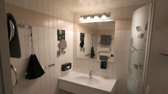 Hvidt flisebelagt badeværelse med vask og spejl, belyst med loftslamper. Metalhåndklædestativ med grå håndklæder. Rundt forstørrelsesspejl på væggen. Matteret glasdør til bruser. Skilte lyder "ÄRON BORG".