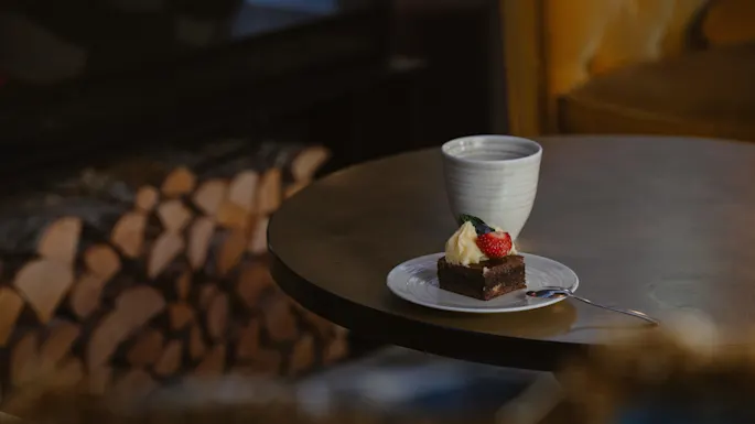 En liten kake med krem og bær ligger på en tallerken ved siden av en kopp på et rundt bord; vedkubber er stablet i bakgrunnen nært en myk stol.