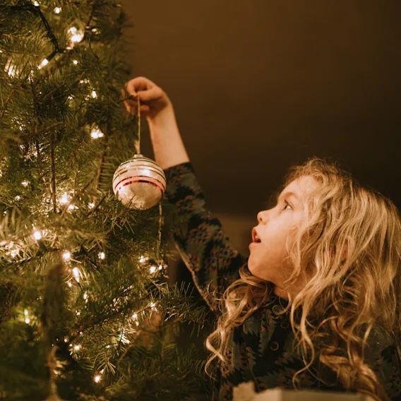 Ei liten jente med krøllete hår henger opp en julekule på et stemningsfullt, lyst juletre.