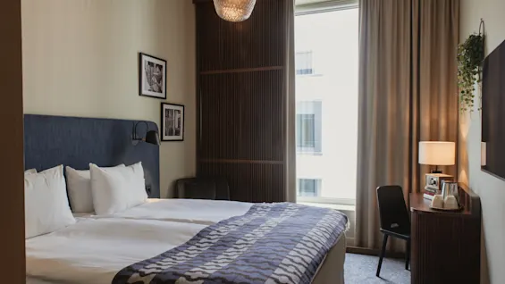Home Hotel Tapetfabriken: A comfortable hotel room with twin beds, a patterned throw, a desk, and a window overlooking the city.