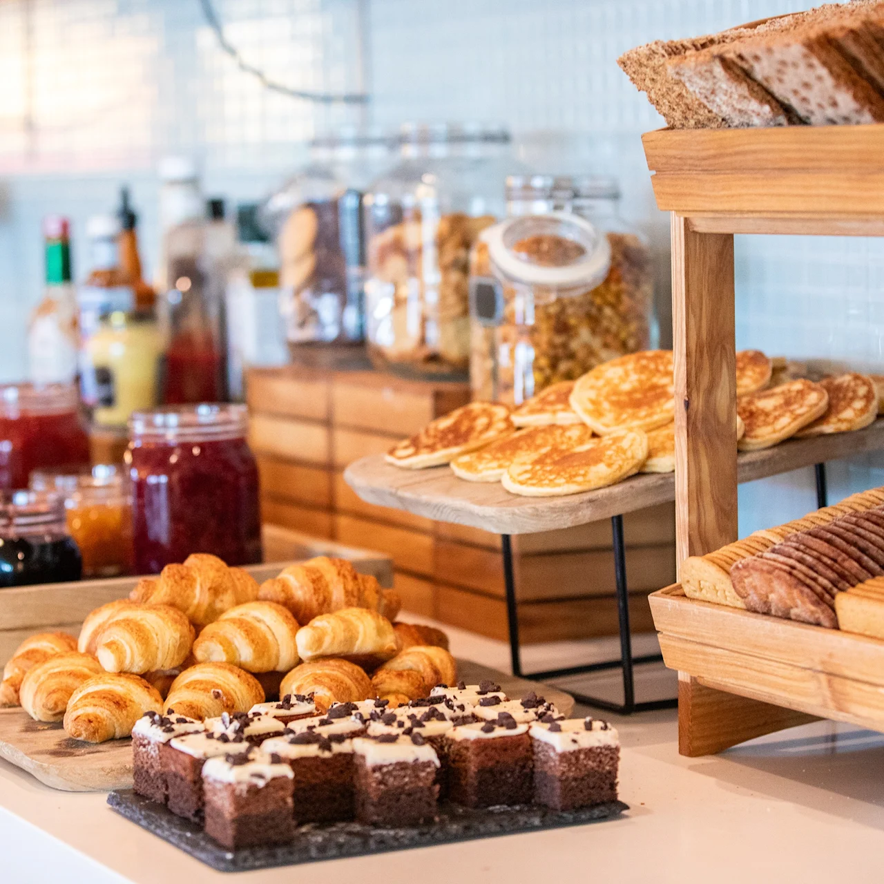 Bakverk og brød er arrangert på et bord, omgitt av syltetøy og frokostblandinger, og skaper en frokostbuffet i et lyst og organisert kjøkkenmiljø.