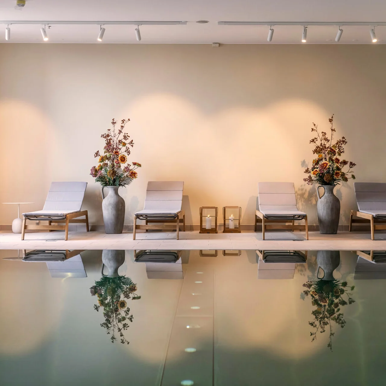 Solstolar uppställda vid en inomhuspool med lanternor och blomvaser. Mjuk belysning kastar reflektioner på vattnet, vilket skapar en lugn spa-liknande atmosfär.