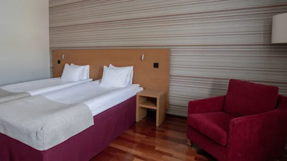 A neatly made twin bed with white linens stands in a room with striped wallpaper. A red armchair sits nearby, and a small wooden nightstand separates the beds.
