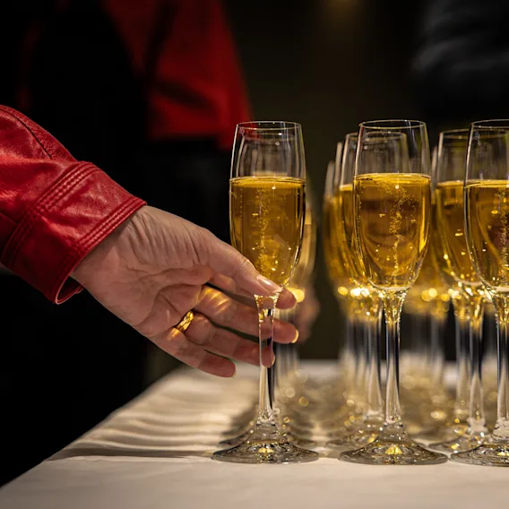 En hand i en röd skinnjacka sträcker sig efter ett glas bubblande champagne från prydligt uppställda rader av fyllda glas på en vit duk.