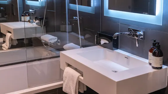 En hvit rektangulær designet vask har et håndkle hengende under. En såpepumpe står på benken. Bak vasken er et badekar synlig med foldede håndklær.