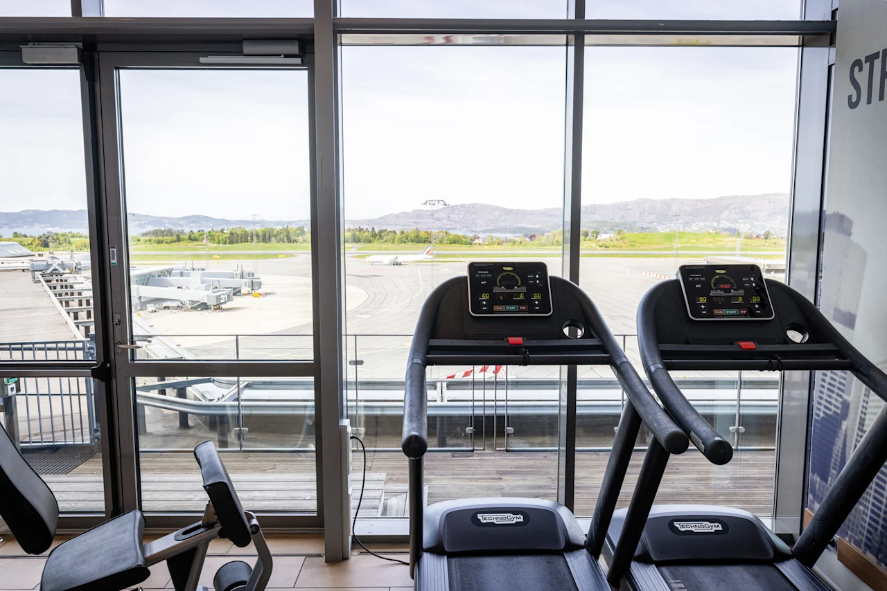 Tredemøller plassert foran store vinduer med utsikt over en flyplass med parkerte fly. Sterkt dagslys belyser treningsområdet. Ingen tekst synlig.