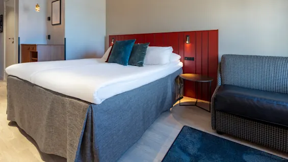A neatly made bed in a modern hotel room with red lockers, a patterned sofa, and warm lighting.