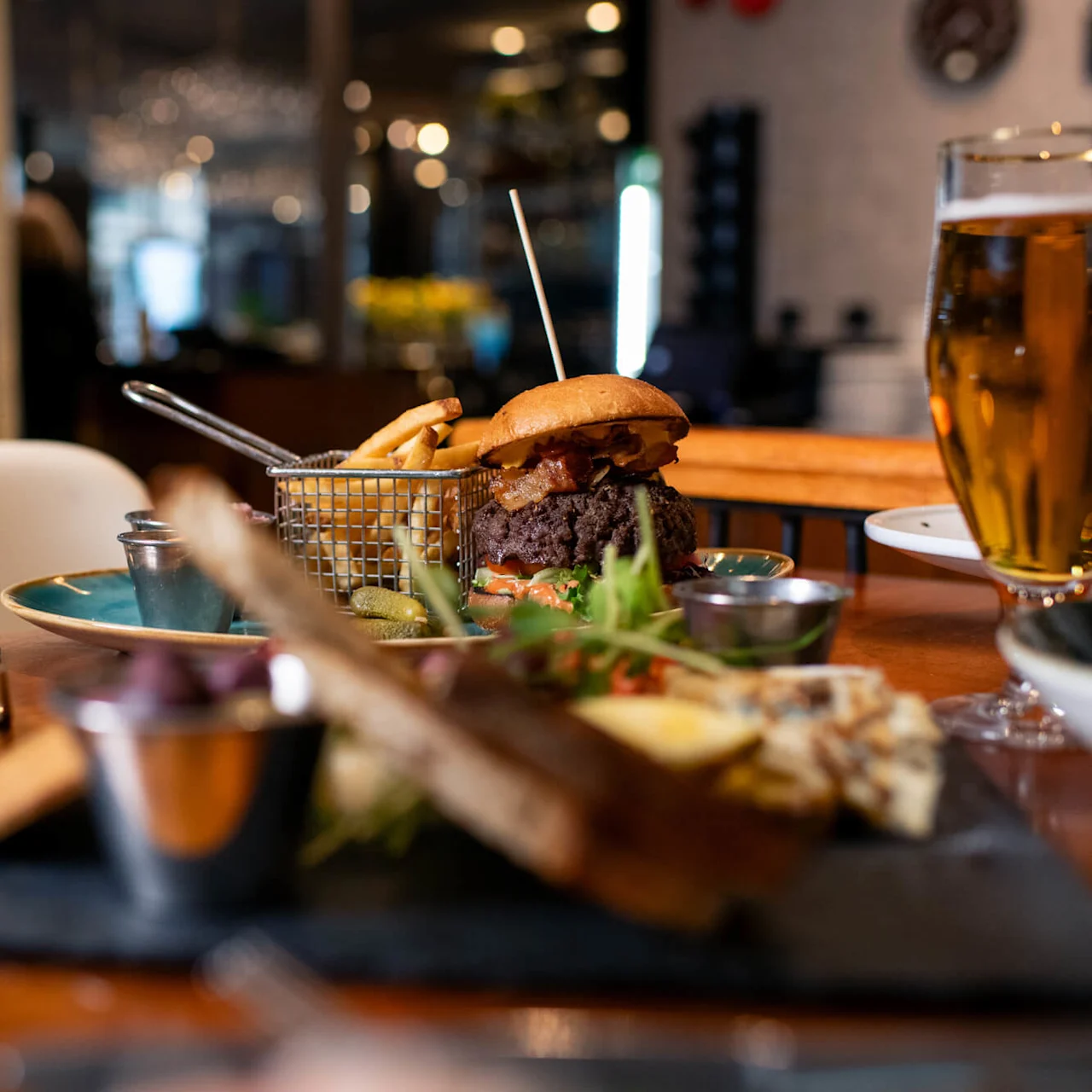Burger sits on a plate with fries in a basket, surrounded by a glass of beer and tableware, in a dimly lit restaurant setting.
