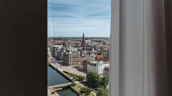 En byutsikt fra et vindu, som viser bygninger, en elv og et kirkespir under en blå himmel.