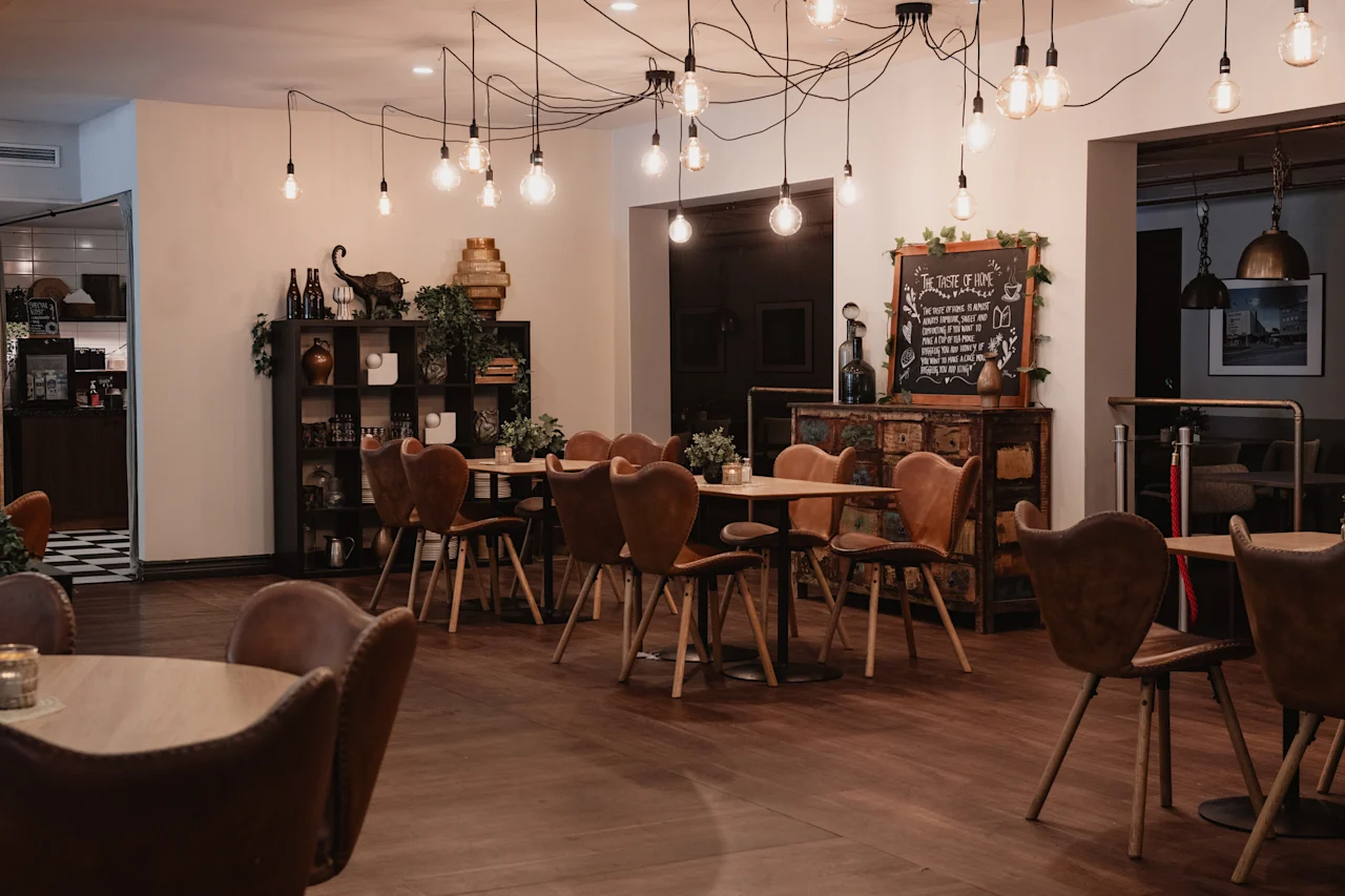 An inviting restaurant or lounge with wooden tables, brown chairs, and exposed light bulbs. A chalkboard sign says THE TASTE OF HOME.