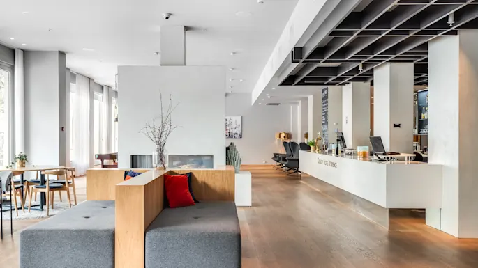 Spacious hotel lobby features a modern sofa with red cushions, a minimalist reception counter, and wooden flooring. Bright natural light streams through large windows. Text on reception: "clarion hotel lumire."