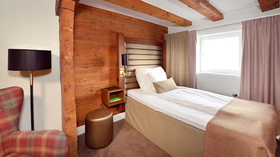 A cozy bedroom with an exposed brick wall, wooden beams, and a single bed with white sheets beside a plaid armchair and a round stool.