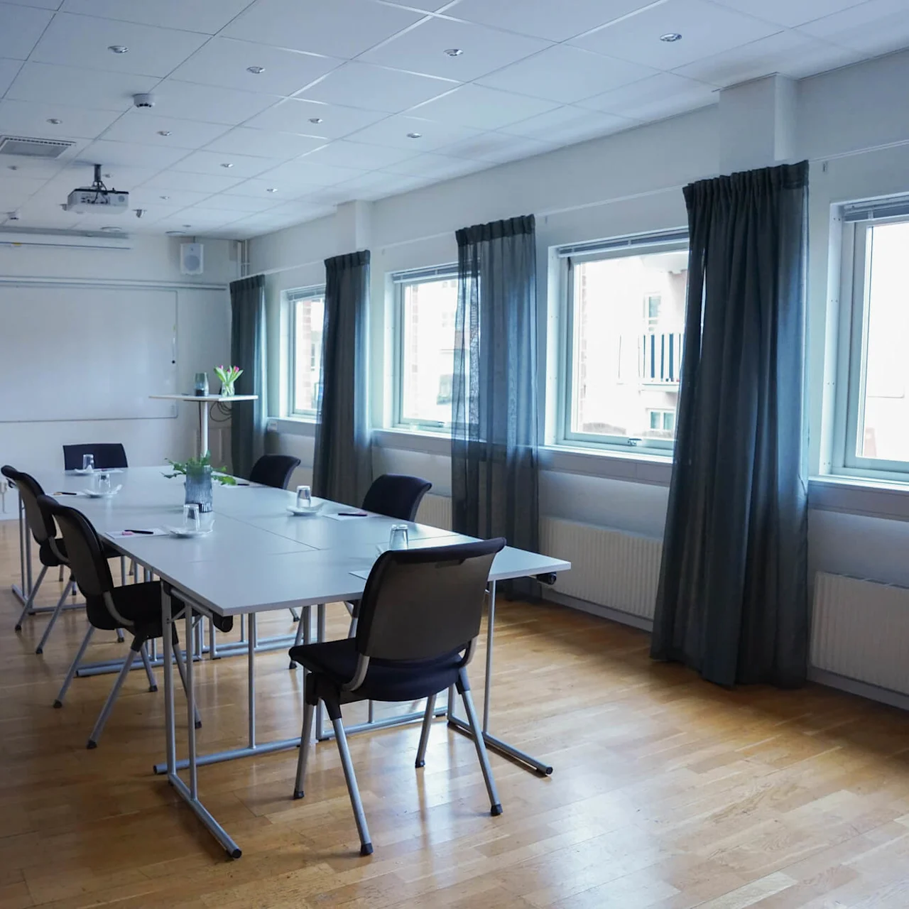 A conference room with a table and chairs, a whiteboard, and a projector, in a well-lit space with windows and curtains.