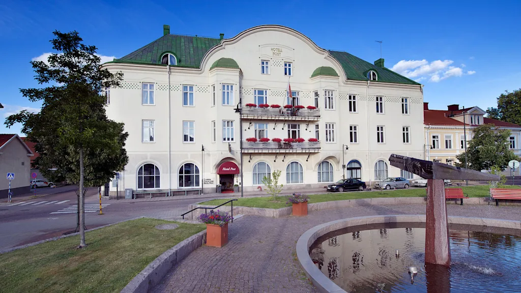 En trestockig byggnad med skylten "HOTELL POST" och balkonger på en solig dag med klar himmel, bredvid en liten damm och parkbänkar.