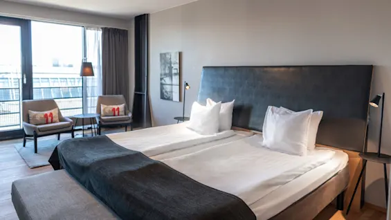 A bed neatly made with white pillows and dark covers stands in a modern hotel room. Two chairs with cushions labeled "11" face a window overlooking buildings.
