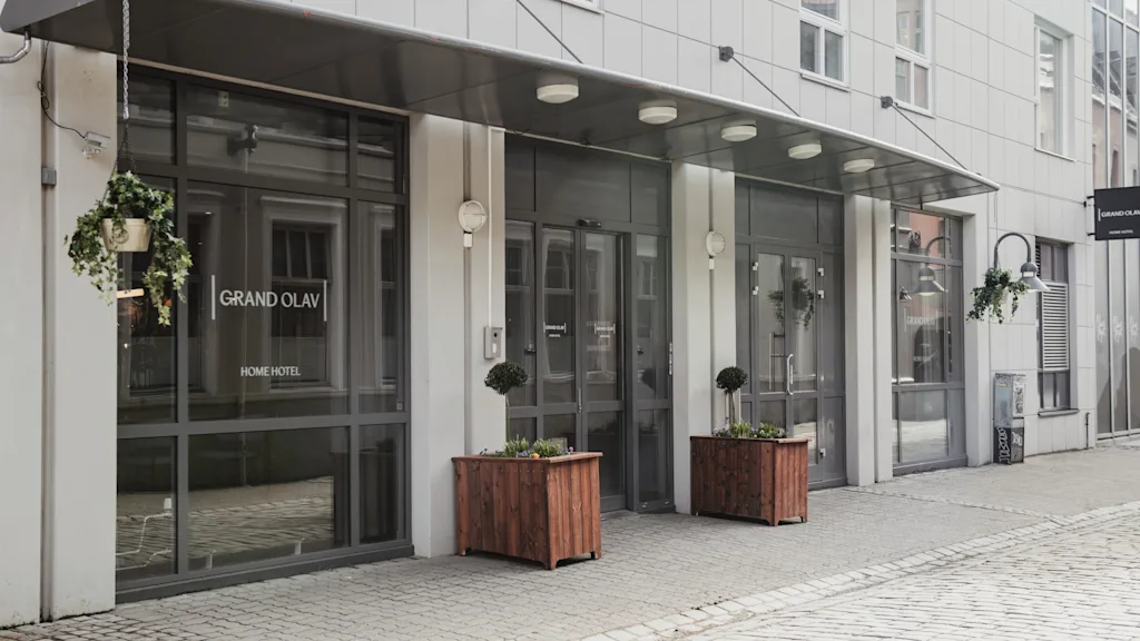 Inngang til en moderne bygning med glassdører, plantekasser og en hengende plante. Tekst på døren lyder GRAND OLAV HOME HOTEL.