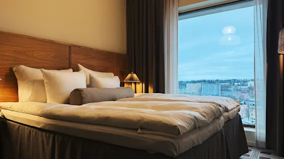 Ett mysigt hotellrum med en stor säng och ett fönster som erbjuder utsikt över en stadssilhuett under en mjuk, molnig himmel.