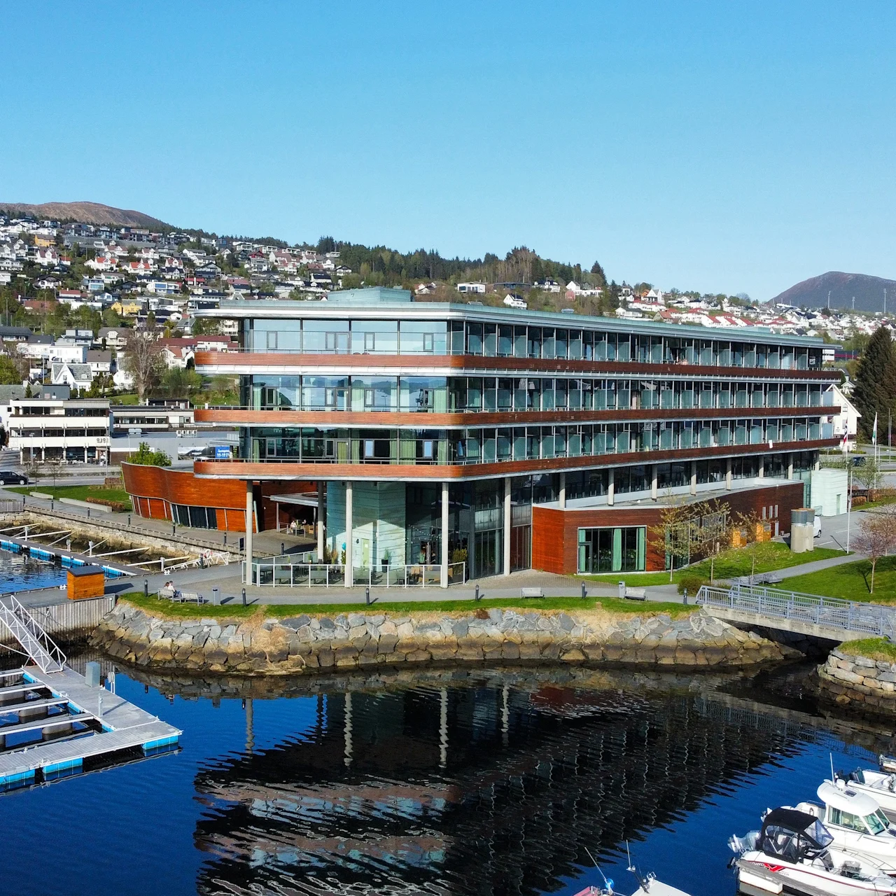 En moderne, fleretasjes bygning med store glassvinduer står ved en marina med flytende brygger. Bolighus og grønne åser er synlige i bakgrunnen.