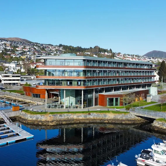En moderne, fleretasjes bygning med store glassvinduer står ved en marina med flytende brygger. Bolighus og grønne åser er synlige i bakgrunnen.