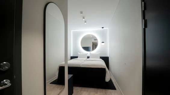A bed with white bedding rests against a wall, beneath a circular, illuminated mirror. The room is minimalist with a wooden floor, and a tall mirror is on the left wall.