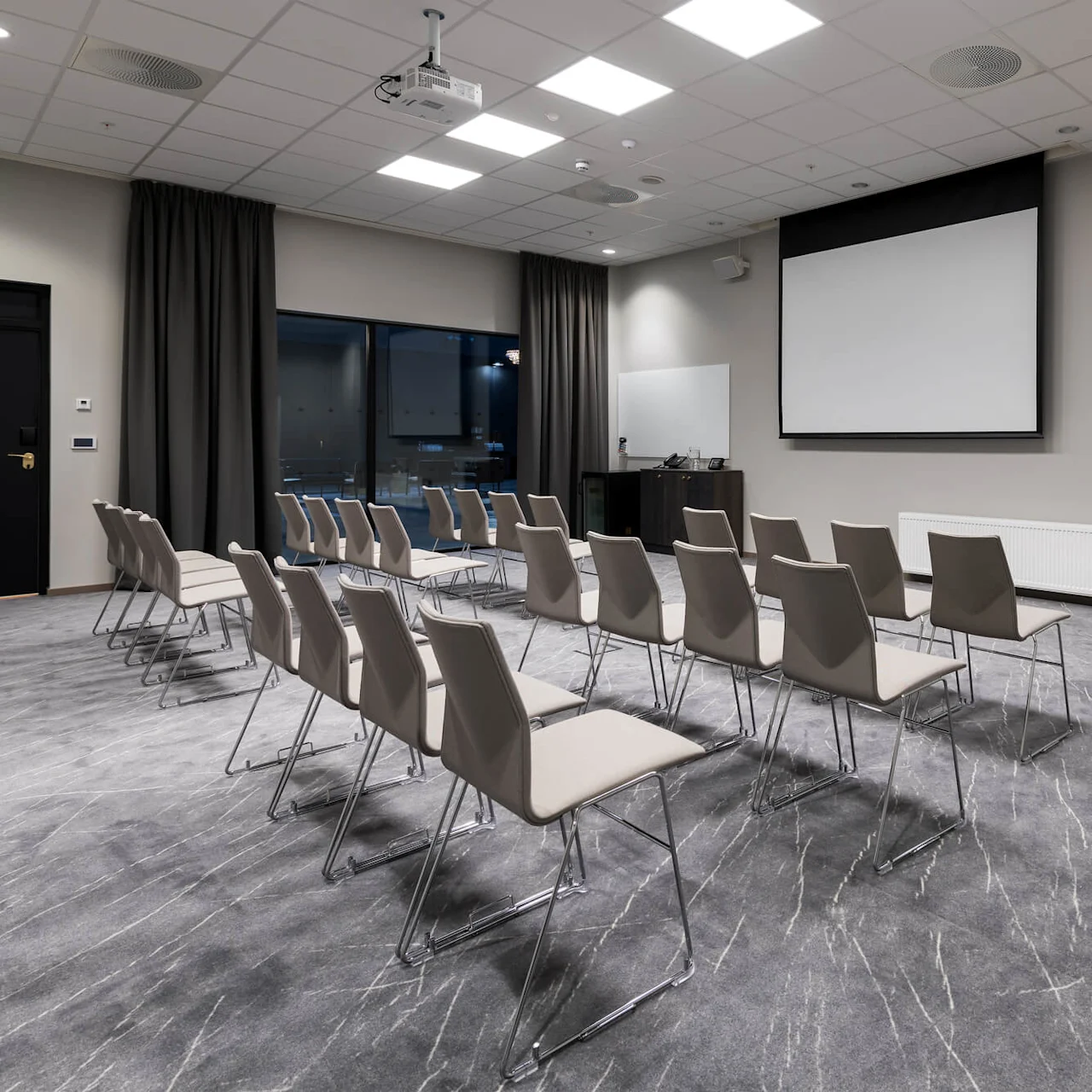 Stoler peker mot en tom projeksjonsskjerm i et godt opplyst konferanserom med grått teppe, et stort vindu og et svart gardin. Ingen tekst er til stede.