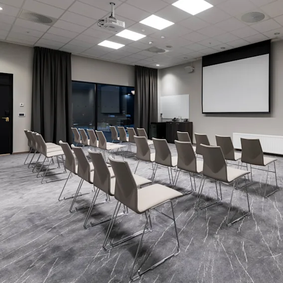 Stoler peker mot en tom projeksjonsskjerm i et godt opplyst konferanserom med grått teppe, et stort vindu og et svart gardin. Ingen tekst er til stede.